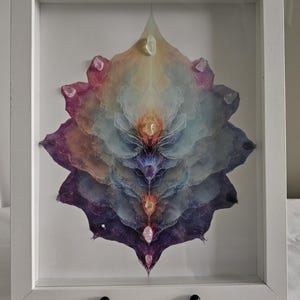 May include: A framed artwork featuring a layered, abstract floral design in shades of purple, blue, yellow, and pink. The artwork is adorned with small, clear crystals and encased in a white wooden frame.
