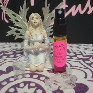 May include: A perfume bottle with a black cap and a pink label that reads "I Am Beautiful Days & Illusions Perfume" sits next to a fairy figurine and a pile of rose quartz crystals. The background is a patterned fabric.