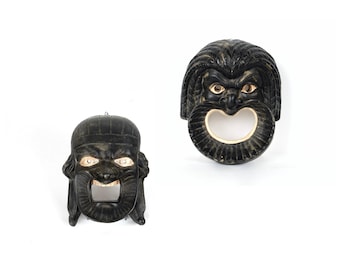 May include: Two black decorative masks with open mouths and detailed facial features. The masks have light-colored eyes and are set against a white background. One mask has a round opening, and the other has a rectangular opening.