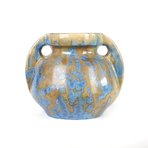 May include: A ceramic vase with a rounded body and two side handles, featuring a glossy glaze with a mottled pattern of tan and blue crystalline spots. The vessel has a wide opening at the top and a smooth, curved silhouette throughout its structure.
