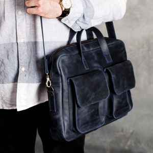 May include: A dark blue leather briefcase with a long strap and two large front pockets. The bag has a zipper closure and a gold metal buckle.