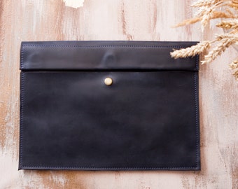May include: A dark blue leather envelope with a brass button closure. The envelope is on a light brown wooden background.