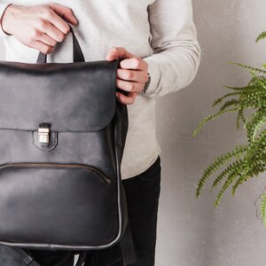 May include: A black leather backpack with a flap closure and a zippered pocket. The backpack has a gold metal buckle and a brown leather strap.