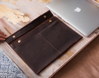 May include: A brown leather laptop sleeve with two pockets and brass snaps. The sleeve is open and a silver laptop is partially visible inside.