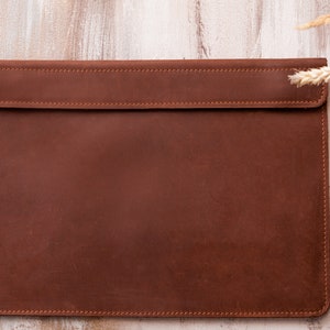Puede incluir: Una funda para portátil de cuero marrón con cierre de solapa y bordes cosidos.