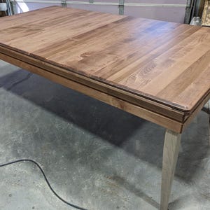 May include: A rectangular dining table featuring a smooth, medium-brown wooden top with visible grain patterns. The table is supported by four tapered, light-colored wooden legs that extend from the corners, providing a stable base structure.