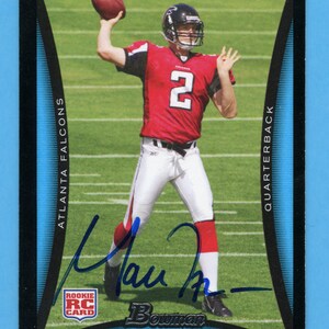 Peut inclure: Carte de recrue de Matt Ryan de Bowman. La carte présente une photo de Ryan dans son uniforme des Falcons d'Atlanta, lançant un ballon de football. La carte a une bordure noire avec des accents bleus et les mots "Atlanta Falcons" et "Quarterback".