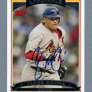 May include: A baseball card featuring Yadier Molina of the St. Louis Cardinals. The card has a white border with red, yellow, and black accents. Molina is wearing a Cardinals uniform and a blue helmet. The card includes the Topps logo.