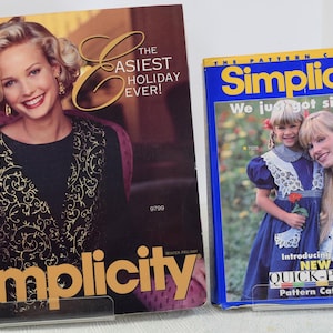 May include: Two Simplicity pattern catalogs. The first catalog features a woman wearing a black top with gold embroidery and the text "The Easiest Holiday Ever!" The second catalog features two girls wearing white lace dresses and the text "We just got simpler! Introducing our New Quick-Pick Pattern Catalog".