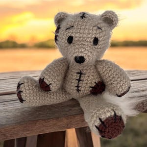 May include: A handmade, light beige crochet teddy bear with dark brown accents on the paws and patches. The bear has black stitched details and is sitting on a wooden surface. The background features a blurred sunset.
