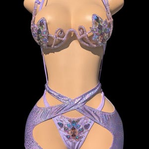 May include: A lavender two-piece outfit with a halter top and shorts. The top features iridescent crystal embellishments and a decorative design. The shorts have cut-out sides and a matching crystal design. The fabric has a subtle, textured pattern.