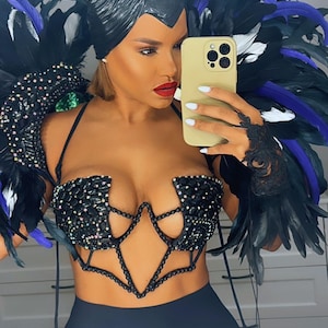 May include: A black costume featuring a horned headpiece, feathered shoulder accents, and a jeweled bodice. The costume includes black and purple feathers, and a black skirt. The person is holding a gold phone.