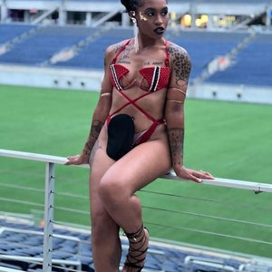 May include: A woman wearing a red and black beaded bra with the text "Parker" and "U.S. Army" on it. She is wearing a red and black beaded bra with a black pouch attached to the front. She is wearing black high-heeled sandals with laces. She is sitting on a railing in front of a stadium.