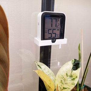 May include: A white digital thermometer and hygrometer with a black display showing 77 degrees Fahrenheit and 55% humidity. The device is mounted on a white plastic bracket.