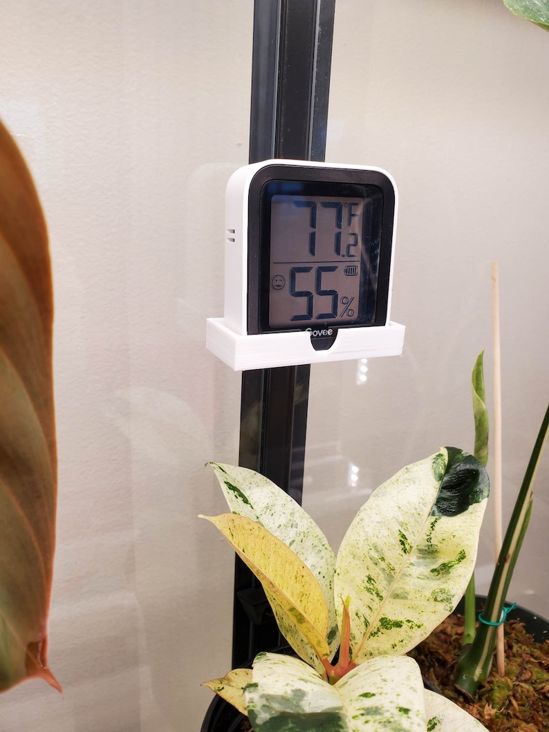 May include: A white digital thermometer and hygrometer with a black display showing 77 degrees Fahrenheit and 55% humidity. The device is mounted on a white plastic bracket.