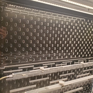May include: A clear acrylic panel with a grid of small, evenly spaced holes. The panel is mounted in a black frame and is illuminated by a row of white LED lights.
