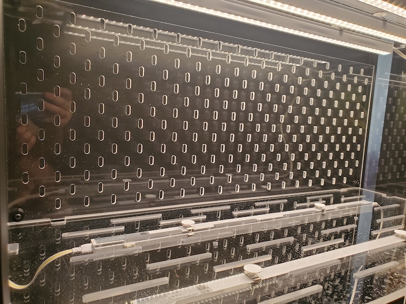 May include: A clear acrylic panel with a grid of small, evenly spaced holes. The panel is mounted in a black frame and is illuminated by a row of white LED lights.