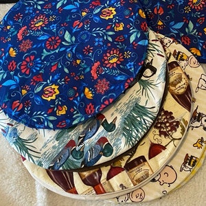 Könnte beinhalten: Vier runde Stoff-Platzsets mit verschiedenen Designs. Das oberste Platzset ist blau mit einem Blumenmuster. Das zweite Platzset ist cremefarben mit einem Enten- und Blatt-Muster. Das dritte Platzset ist cremefarben mit einem Trauben- und Weinflaschen-Muster. Das unterste Platzset ist cremefarben mit einem Cartoon-Hund und anderen Cartoon-Charakteren.