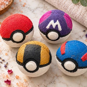 May include: Four spherical bath bombs designed to resemble various ball-shaped items. One is red and white, one is purple and white with a white letter M, one is gold and black, and one is blue and white with red accents. Each has a central white circle.