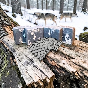 Puede incluir: Jabones artesanales con un diseño veteado en gris y blanco, junto con un emblema de cabeza de lobo gris. Los artículos están dispuestos sobre una superficie de madera envejecida, con lobos en el fondo nevado. Los jabones miden unos 7 cm de largo.