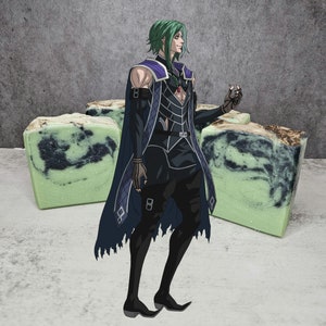 Puede incluir: Un personaje con cabello verde y una capa de color morado oscuro se encuentra frente a dos barras de jabón verde y negro. El personaje lleva un atuendo negro y plateado con una joya roja en el pecho. El personaje sostiene un objeto pequeño en su mano derecha.