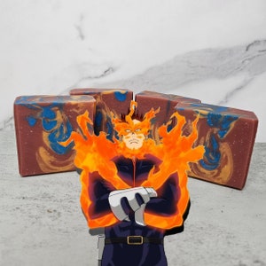 Puede incluir: Una pastilla de jabón hecha a mano con un patrón de remolino de azul, marrón y dorado. El jabón tiene forma de rectángulo y está sobre una superficie blanca. Un personaje de dibujos animados con cabello naranja y un atuendo azul y negro está frente al jabón.