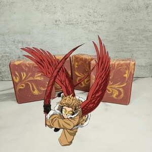 Puede incluir: Una pastilla de jabón roja y dorada con una imagen estilizada de un personaje alado con cabello rubio y una chaqueta marrón. El personaje sostiene una pluma roja y dorada. La pastilla de jabón está sobre una superficie blanca.