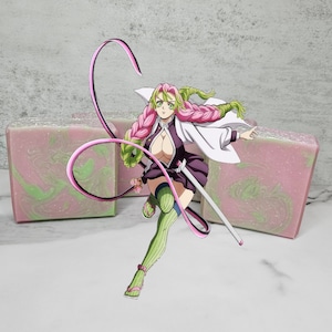 Puede incluir: Una barra de jabón de color rosa y verde con un personaje del anime Demon Slayer. El personaje es una mujer con cabello rosa y ojos verdes, vestida con un atuendo blanco y morado. Ella sostiene una espada y tiene una cinta rosa y verde atada alrededor de su cintura.