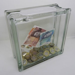 May include: A clear glass block money bank with a white plastic slot. Inside are banknotes and coins. The banknotes feature Queen Elizabeth II. The coins are gold and silver. A transparent money box for saving.