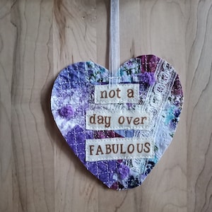 May include: A purple and white patchwork heart-shaped ornament with the text "not a day over FABULOUS" hanging from a white ribbon.