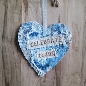 May include: A blue and white fabric heart-shaped ornament with the words "CELEBRATE today" stitched on it. The ornament is hanging from a white ribbon.