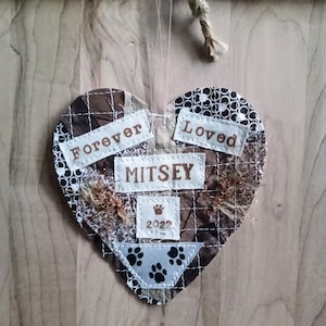 May include: A brown and white patchwork heart-shaped ornament with the words "Forever Loved" and the name "Mitsey" on it. The ornament also has a paw print and the year "2022" on it.
