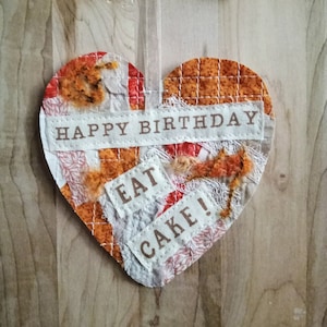 May include: A heart-shaped fabric craft with a brown and orange patchwork design. The fabric has a distressed look with white lace and fabric scraps. The text "Happy Birthday Eat Cake!" is printed on the fabric.