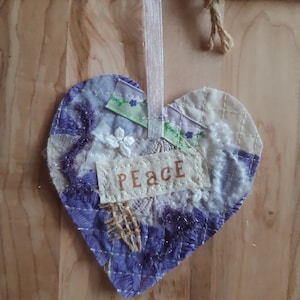 May include: A purple and white heart-shaped fabric ornament with the word "Peace" written on it. The ornament is decorated with lace, ribbon, and other fabric scraps.