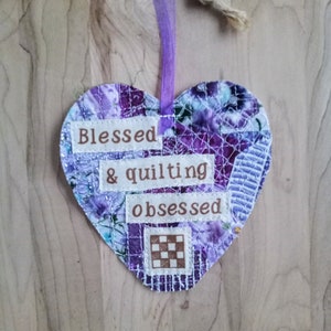 May include: Purple and white heart-shaped fabric ornament with the text "Blessed & quilting obsessed" on it. The ornament is hanging from a purple ribbon.