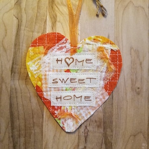 May include: A heart-shaped fabric ornament with a patchwork design in orange, white, and brown. The ornament has the words "HOME SWEET HOME" stitched on it.