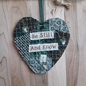 May include: Heart-shaped fabric ornament with a patchwork design in shades of green and gray. The ornament features the text "Be Still And Know" in white lettering. A green ribbon and twine are attached for hanging.