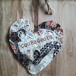 May include: A brown, white, and black patchwork heart-shaped ornament with a white lace trim and a brown ribbon. The ornament has a white tag with the text "Courageous" in brown lettering.