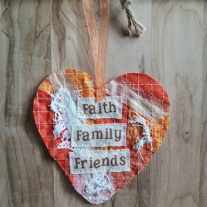 May include: A heart-shaped fabric ornament with the words "Faith", "Family", and "Friends" stitched on it. The ornament is made of patchwork fabric in shades of orange, red, and white.