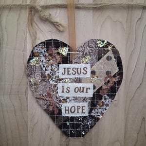 May include: Heart-shaped wall hanging with the text "JESUS is our HOPE." The heart is made of fabric patches in brown, white, and gold tones, with a grid overlay. A tan ribbon is attached for hanging.