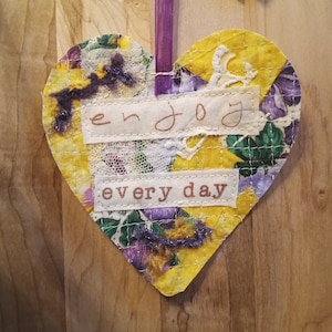 May include: A yellow heart-shaped fabric ornament with purple and white accents. The ornament has the words "enjoy every day" written on it.