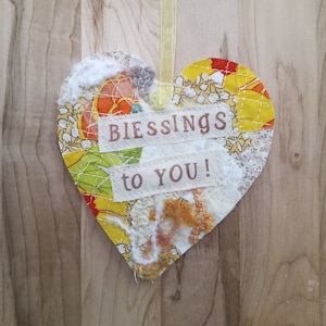 May include: A heart-shaped fabric ornament with a patchwork design in yellow, orange, white, and green. The ornament has two white fabric labels with the text "Blessings to You!" in brown lettering.
