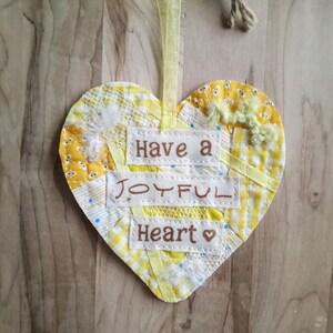 May include: A yellow heart-shaped fabric ornament with the words "Have a Joyful Heart" embroidered on it.