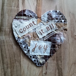 May include: A heart-shaped fabric ornament with a patchwork design. The ornament is decorated with brown, white, and gold fabric scraps and features the words "count it all joy" in brown lettering.