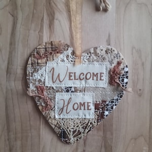 May include: A heart-shaped fabric wall hanging with a patchwork design in shades of brown and beige. The hanging features the words "Welcome Home" in a cursive font.