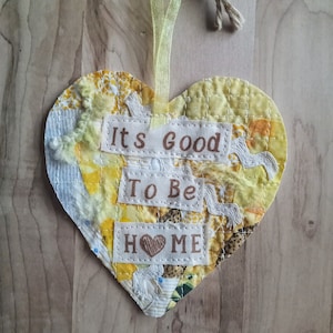 May include: A heart-shaped fabric ornament with the words "It's Good To Be Home" stitched on it. The ornament is made of various yellow and white fabrics and has a rustic, handmade look.
