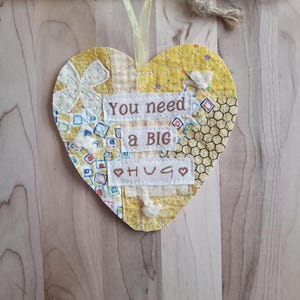 May include: A handmade, yellow fabric heart with the text "You need a BIG HUG" embroidered on it. The heart is decorated with various fabric patterns and a yellow ribbon for hanging. The background is a light wood grain.