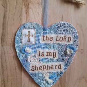 May include: A blue heart-shaped fabric ornament with the words "the LORD is my Shepherd" and a cross. The ornament is decorated with lace and other fabric scraps.
