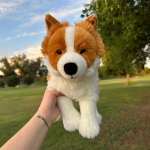 May include: A plush toy dog with a white and brown coat, black nose, and soft, fluffy fur. The stuffed animal is held up against a background of green grass and a blue sky with clouds.