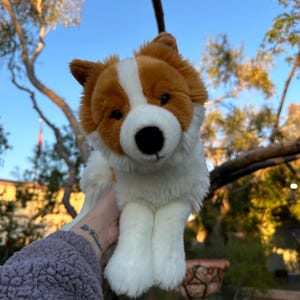 May include: A plush dog toy with a white body, brown ears, and a brown and white face. The toy has a black nose and is held up against a blue sky with tree branches in the background.